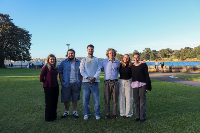 A group of young people stand together on grass with the harbour in the backdrop